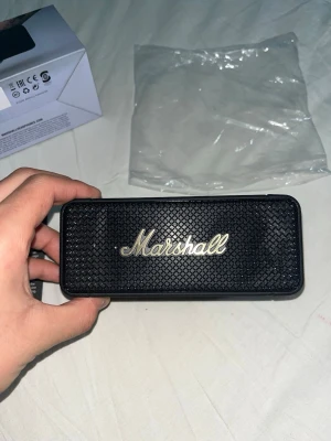 Marshall svart bärbar högtalare - Stilren bärbar högtalare från Marshall i svart med klassiskt metallgaller och Marshall-logga i guld. Högtalaren har en rektangulär form, gummerad yta och detaljer i metall. På ovansidan finns volymratt, knappar för ström och volym samt laddningsport.
