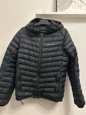 Svart pufferjacka från Peak Performance - Säljer en svart pufferjacka från Peak Performance med huva och dragkedja framtill. Jackan har diskret logga på bröstet och är lätt vadderad med horisontella sömmar. Perfekt för kalla dagar och enkel att matcha med olika outfits.