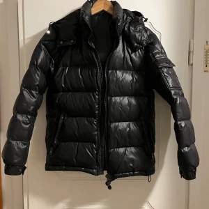 Svart dunjacka med huva - Snygg svart dunjacka,  liten S,med vadderad huva och dragkedja framtill. Jackan har två sidofickor med dragkedja och quiltad design som ger en puffig look. Perfekt för kalla vinterdagar och håller dig varm med sin tjocka stoppning.nyskick. Kan passa både kille/tjej. (Nypris 1100)
