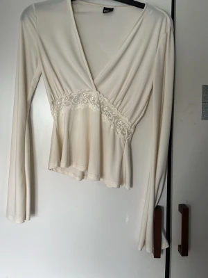 Vit omlottopp med spetsdetalj - Snygg vit omlottopp med v-ringning och långa ärmar. Toppen har en vacker spetsdetalj med blommönster under bysten och lätt utsvängd nederdel. Perfekt för dig som gillar romantisk och feminin stil.