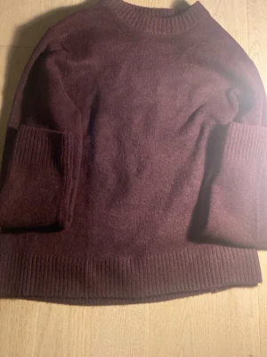 Mörklila stickad tröja med ribbade muddar - Mysig vinröd  stickad tröja med rund halsringning och ribbade muddar vid ärmslut och nederkant. Tröjan har en enkel och stilren design, perfekt för lager-på-lager och chill dagar. Materialet ser ut att vara mjukt och skönt.