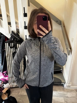 Grå sportig hoodie med dragkedja - Snygg gråmelerad hoodie med hel dragkedja och huva. Jackan har svarta detaljer, två fickor med dragkedja och diskreta reflexprickar vid fickorna. Perfekt för träning eller chill. Mjukt och stretchigt material.