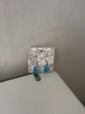 Blå björngodis örhängen - Säljer ett par unika örhängen med blå björngodis-berlocker i plast. Örhängena har silvriga krokar och en lekfull design som verkligen sticker ut. Perfekta för dig som gillar färgglada accessoarer och vill ha något annorlunda i smyckessamlingen.