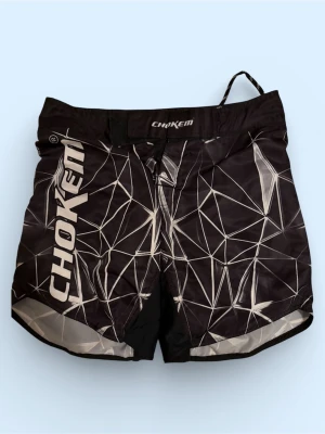 MMA shorts från Chokem - Snygga svarta träningsshorts från Chokem med vitt geometriskt mönster och stor logga på både fram- och baksida. Shortsen har snörning i midjan och är tillverkade i ett lätt syntetmaterial som passar perfekt för intensiva träningspass.