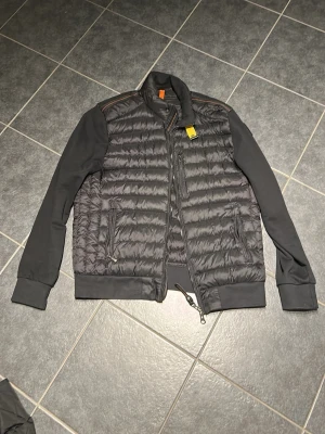 Svart pufferjacka från Parajumpers - Snygg svart pufferjacka från Parajumpers med quiltad front och släta ärmar. Jackan har dragkedja, ståkrage och ikonisk PJS-patch på ärmen. Perfekt för dig som vill ha en stilren och sportig look. Passar till det mesta och är riktigt bekväm.