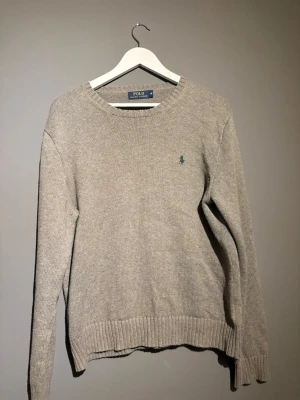 Grå stickad tröja Polo Ralph Lauren - Klassisk grå stickad tröja från Polo Ralph Lauren med rund halsringning och ribbade muddar. Tröjan har den ikoniska gröna logotypen broderad på bröstet. Perfekt för lager på lager och enkel att matcha med olika stilar.