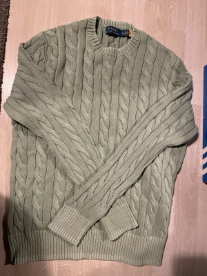 Grön kabelstickad tröja Polo Ralph Lauren - Snygg ljusgrön kabelstickad tröja från Polo Ralph Lauren. Tröjan har rund halsringning, ribbade muddar och nederkant samt ett klassiskt flätmönster. Perfekt för dig som gillar stilrena och tidlösa plagg i mjukt material. Skriv vid frågor 