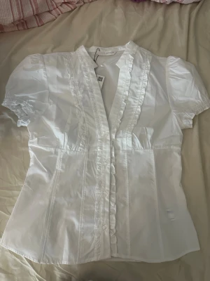 Vit blus med volangkant från Zara  - Söt vit blus från Zara med korta puffärmar och volangkant längs knäppningen. Blusen har v-ringning och är tillverkad i ett lätt, vävt material som känns luftigt. Perfekt att styla med jeans eller kjol för en fräsch look.