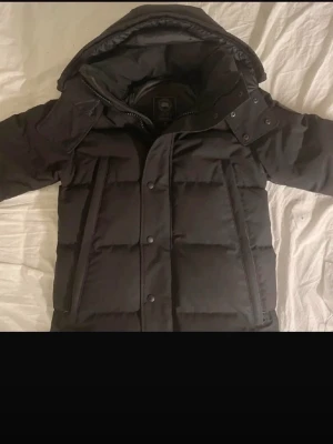 Svart dunjacka från Canada Goose - Säljer en svart dunjacka från Canada Goose med hög krage och avtagbar huva. Jackan har två stora dragkedjeförsedda fickor framtill och klassisk Canada Goose-logga på ärmen. Perfekt för kalla vinterdagar tack vare det tjocka dunfodret.