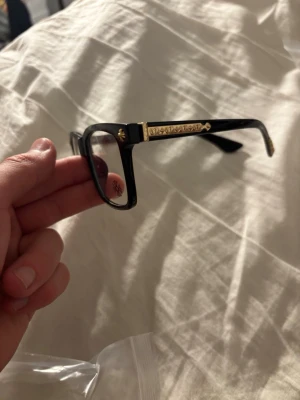 Chrome heart glasögon  - Snygga glasögon med svart båge och rektangulär form. Sidorna har detaljer i guld med mönster och små nitar för en lyxig känsla. På glaset finns en dekorativ logga. Perfekt accessoar för att lyfta din stil.