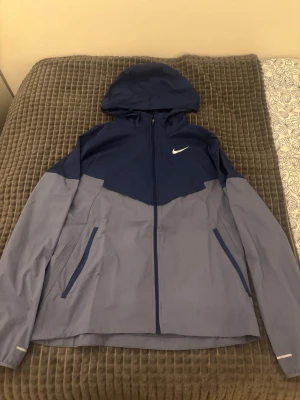 Nike tvåfärgad vindjacka med huva - Nike vindjacka i blått och grått med huva och dragkedja framtill. Jackan har två sidofickor med dragkedja och reflekterande detaljer på ärmar och rygg. Tillverkad i lätt och smidigt material, perfekt för blåsiga dagar. Nike-logga på bröstet.                      DET FINNS EN LITEN FLÄCK SOM TYP LIGGER LÄNGST NER LÅ FRAMSIDAN KNAPPATS SYNLIG PÅ VÄNSTER SIDA!
