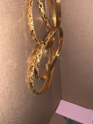 Guldiga armband med gravyr - Säljer ett set med fyra guldiga armband i metall. Armbanden har en rund form och är dekorerade med detaljerade, ingraverade mönster som ger en lyxig och unik känsla. Perfekta för att lyfta vilken outfit som helst med lite extra bling.