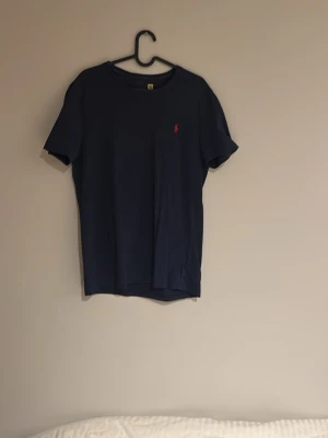 Mörkblå t-shirt från Polo Ralph Lauren - Snygg mörkblå t-shirt från Polo Ralph Lauren med klassisk röd broderad logga på bröstet. T-shirten har rund hals och korta ärmar. Perfekt basplagg med stilren design och mjukt material.