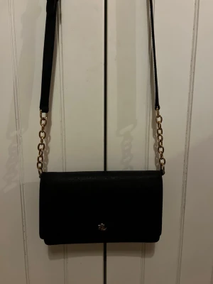 Svart väska från lauren Ralph Lauren - Snygg svart axelväska från Ralph Lauren med guldfärgade detaljer och kedja på axelremmen. Väskan har ett stilrent LRL-emblem framtill och magnetlås. Äkta!!