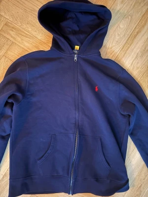 Mörkblå hoodie från Polo Ralph Lauren - Mörkblå hoodie med dragkedja från Polo Ralph Lauren. Klassisk design med huva, två fickor framtill och den ikoniska röda logotypen broderad på bröstet. Storlek 170-176. Tillverkad i mjukt bomullsmaterial som är skönt att ha på sig. Pris ej hugget i sten!