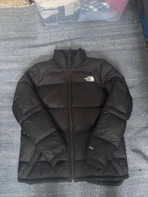 Svart dunjacka från The North Face - Säljer en svart dunjacka från The North Face med klassisk logga på bröstet. Jackan har hög krage, dragkedja framtill och quiltad design. Perfekt för kalla dagar och enkel att matcha med olika outfits.