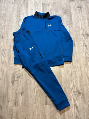 Under Armour - Snyggt blått träningsset från Under Armour med matchande tröja och byxor. Tröjan har halv dragkedja, svart krage och vit logga på bröstet. Byxorna har resår i midjan, dragkedjefickor och vit logga på låret. Perfekt för träning eller chill.