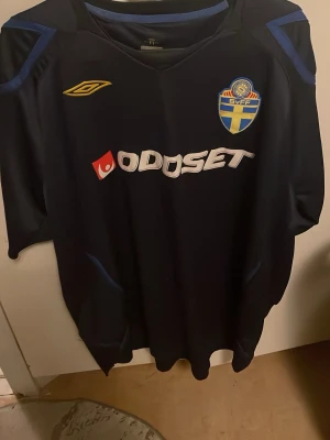 Svenska landslagströja Umbro blå - Mörkblå fotbollströja från Umbro med Svenska Fotbollsförbundets märke på bröstet och Oddset-logga framtill. Tröjan har gula och blå detaljer och är tillverkad i ett lätt och ventilerande material, perfekt för träning eller match.
