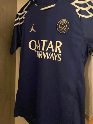 Blå PSG matchtröja D.Doué #14 - Snygg blå PSG matchtröja med vita detaljer och vingmönster på axlarna. Framsidan har Paris Saint-Germain-logga, Jordan-logga och Qatar Airways-tryck. Baksidan har D.Doué och nummer 14. Perfekt för fotbollsfans och samlare.