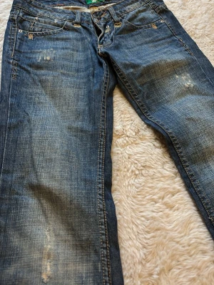 Blå bootcut low waist jeans - Snygga blå bootcut jeans från Bershka med slitna detaljer och ljusare partier framtill. Klassisk femficksmodell med låg midja och knappgylf. Perfekta för en avslappnad och trendig look.