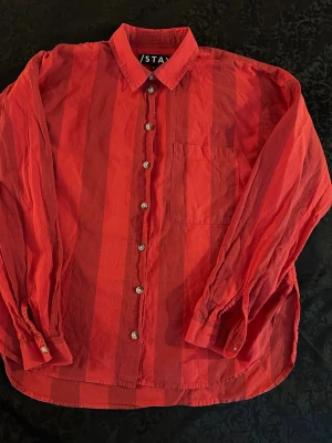 Röd randig skjorta från Stay - Säljer en röd skjorta från Stay med breda vertikala ränder och klassisk krage. Skjortan har knappar framtill, en bröstficka och långa ärmar. Perfekt för dig som gillar färg och vill sticka ut lite extra.