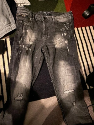 Svarta distressed jeans Supply & Demand - Säljer ett par svarta jeans från Supply & Demand med slitna detaljer, blekta partier och dragkedjeficka fram. Jeansen har skinny fit och är tillverkade i denim med stretch. Perfekta för en edgy streetwear-look.