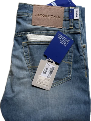 Jacob Cohën Jeans (Nya med Tags) | W31 - Snygga ljusblå jeans från Jacob Cohën, helt nya med tags. Ordinarie nypris ca 3800. Klassisk femficksmodell och raka ben. Jeansen har en stor läderpatch bak i midjan och kontrastsömmar. Tillverkade i mjuk denim med knappgylf och diskret tvättad look.