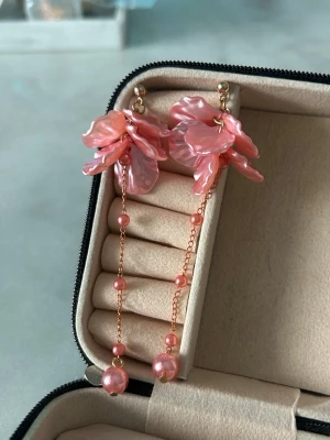 Rosa blomformade örhängen med pärlor - Unika örhängen i form av rosa blommor med skimrande yta och hängande kedjor dekorerade med små rosa pärlor. Örhängena har en elegant och lekfull design som verkligen sticker ut. Perfekta för dig som vill ha något extra till din stil.