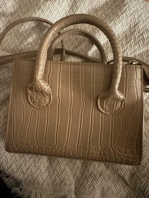 Beige handväska med krokopräglat mönster - Snygg beige handväska i fyrkantig modell med krokopräglat mönster. Väskan har både handtag och axelrem samt dragkedja upptill. Perfekt för dig som gillar stilrena accessoarer med lite extra detalj.