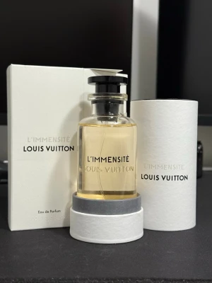 Louis Vuitton L'Immensité Eau de Parfum 100ml - Exklusiv parfym från Louis Vuitton, L'Immensité, i elegant glasflaska med svart lock. Doften kommer i en stilren vit förpackning och cylinderetui. Flaskan rymmer 100 ml och har en lyxig, minimalistisk design som utstrålar premiumkänsla.