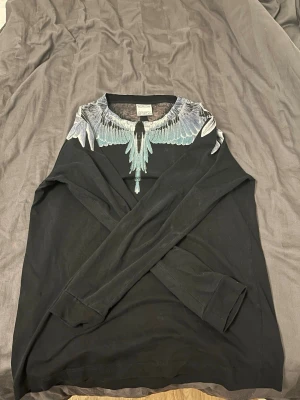 Svart tröja från Marcelo Burlon - Svart långärmad tröja från Marcelo Burlon med ikoniskt vingeprint i grått och turkost runt halsen. Tröjan har en avslappnad passform och är tillverkad i mjukt bomullsmaterial. Perfekt för dig som gillar streetwear och unika detaljer.