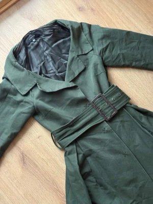 Mörkgrön trenchcoat med bälte från Zara - Stilren mörkgrön trenchcoat med klassisk krage, knäppning framtill och matchande bälte i midjan. Jackan har slät insida och långa ärmar, perfekt för en trendig look. Materialet är mjukt och följsamt, vilket ger en snygg siluett. Har även fickor. Knapparna kan behövas sys på lite bättre men funkat fortfarande.