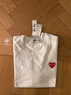 Vit Comme des Garçons Play t-shirt - Vit t-shirt från Comme des Garçons Play med det ikoniska röda hjärtat med ögon på bröstet och ett stort hjärtmotiv längst ner. Tröjan är helt ny utan defekter och är i storlek S