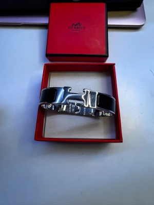 Armband  - Snyggt svart armband från Heeermessss med ikoniskt H-spänne i silverfärgad metall. Armbandet har en stilren och modern design med blanka svarta partier och kommer i original ask. Perfekt accessoar för att lyfta din outfit. Jage öppen för bud.