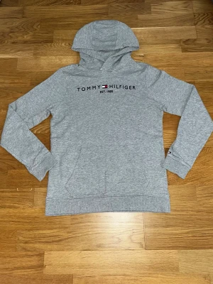 Grå hoodie från Tommy Hilfiger STRl:164 - Snygg grå hoodie från Tommy Hilfiger med broderad logga och text 'EST. 1985' på bröstet. Klassisk känguruficka framtill och huva. Tillverkad i mjukt bomullsmaterial som känns skönt mot huden. Perfekt för en avslappnad och stilren look.