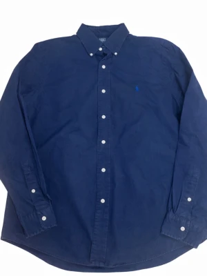 Marinblå skjorta från Ralph Lauren - Klassisk marinblå skjorta från Ralph Lauren med button-down krage och vita knappar. Skjortan har lång ärm och den ikoniska broderade loggan på bröstet. Tillverkad i mjuk bomull som känns skön mot huden. Perfekt för en clean och stilren look.