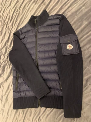 Mörkblå cardiganjacka från Moncler - Snygg mörkblå cardiganjacka från Moncler med quiltad front och stickade ärmar. Jackan har dragkedja framtill, ribbade muddar och en ficka med Moncler-logga på ärmen. Perfekt för lager-på-lager och streetwear-stil.