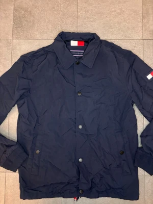 Marinblå vindjacka Tommy Hilfiger - Stilren marinblå vindjacka från Tommy Hilfiger med klassisk krage, tryckknappar framtill och två sidofickor. Jackan har diskret logga på ärmen och är tillverkad i ett lätt, vindtätt material som passar perfekt till svala dagar.