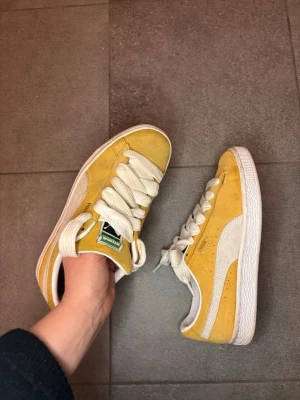 Gula Puma Suede sneakers - Säljer ett par gula Puma Suede sneakers med vit sula och vita detaljer. Skorna har breda vita skosnören och den klassiska Puma-loggan på sidan. Ovandelen är i mocka och modellen är låg. Perfekta för dig som vill sticka ut med färg!