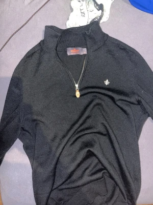 Svart stickad tröja från Morris - Snygg svart stickad tröja från Morris med halv dragkedja och ståkrage. Tröjan har en liten broderad logga på bröstet och ribbade muddar. Perfekt för dig som gillar stilrena plagg med diskreta detaljer.