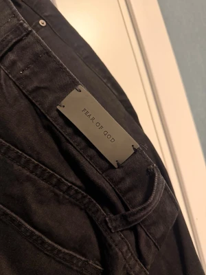 Svarta Fear of God jeans med slitningar - Säljer ett par svarta jeans från Fear of God med skinny fit och slitningar vid knäna. Klassisk femficksmodell med metallknappar och detaljer. Perfekta för dig som gillar streetwear och vill ha en edgy look.