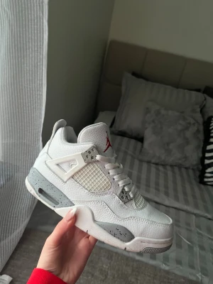 Air Jordan 4 White Oreo sneakers - Säljer ett par Air Jordan 4 White Oreo sneakers med vit ovandel i läder, mesh-detaljer och grå sula med svarta stänk. Klassisk siluett med röda detaljer på plösen och ikoniska plastdetaljer vid snörningen. Perfekta för dig som gillar stilrena och sportiga sneakers.