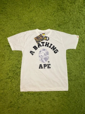 Vit A Bathing Ape t-shirt med tryck - Säljer en vit t-shirt från A Bathing Ape med det klassiska BAPE-logotypen och texten i svart på bröstet. T-shirten har rund hals, korta ärmar och är gjord i mjuk bomull. Perfekt för dig som gillar streetwear och vill sticka ut med en ikonisk design.