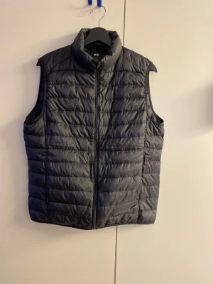 Svart dunväst från Uniqlo - Snygg svart dunväst från Uniqlo med lättviktigt material och vattenavvisande yta. Västen har dragkedja framtill, hög krage och quiltad design. Perfekt för lager-på-lager och enkel att matcha med olika outfits.