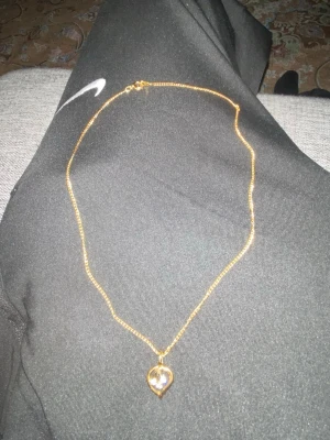 Guldigt halsband med hjärthänge - Snyggt halsband i guldfärg med ett hjärtformat hänge. Hänget har en glittrande sten i mitten som ger extra bling. Kedjan är tunn och elegant, perfekt för att lyfta vilken outfit som helst. Märke är okänt men det ör guld!