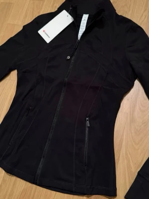 Svart Luon Define träningsjacka från Lululemon - Helt ny snygg svart Luon träningsjacka från Lululemon med figurnära passform, hel dragkedja framtill och två fickor med dragkedja. Prislapp kvar på! Tillverkad i ett stretchigt funktionsmaterial som passar perfekt till träning eller som ett extra lager. Diskret logga på dragkedjan och på baksidan av nacken. Skickas självklart i originalförpackningen!💖💕