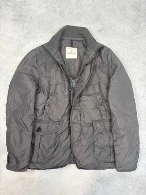 Grå pufferjacka från Moncler - Snygg grå pufferjacka från Moncler med hög krage i ribbad bomull, dragkedja och knappar framtill. Jackan har två stora fickor med lock och knapp samt två bröstfickor. Tillverkad i glansigt, quiltat material som håller värmen under kalla dagar.