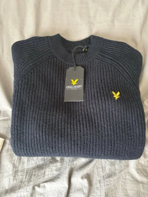 Mörkblå stickad tröja Lyle & Scott - Helt ny mörkblå stickad tröja från Lyle & Scott med klassisk rund halsringning och ribbstickad struktur. Tröjan har den ikoniska gula logon broderad på bröstet och är tillverkad i ett mjukt material som passar perfekt till kyligare dagar. 599 kr! Inköpt för 999 nedsänkt från 1549! Prislapp kvar! Kan gå ner lite i pris vid enkel och snabb affär!