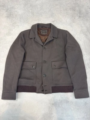 Brun jacka från Canali med knappar - Snygg brun jacka från Canali med klassisk krage och knäppning framtill. Jackan har två stora fickor med lock och knappar samt ribbade muddar vid ärmslut och nederkant. Tillverkad i ett strukturerat tyg som ger en exklusiv känsla.