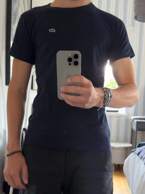 Mörkblå Lacoste t-shirt i bomull - Snygg mörkblå t-shirt från Lacoste med klassisk krokodillogo på bröstet. T-shirten har rund halsringning och korta ärmar. Tillverkad i mjuk bomull som känns skön mot huden. Perfekt för en clean och stilren look. Storlek 14 (164cm)